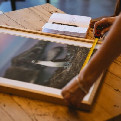 Person measuring a framed photograph with a tape measure on a wooden table indoors.