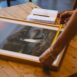 Person measuring a framed photograph with a tape measure on a wooden table indoors.